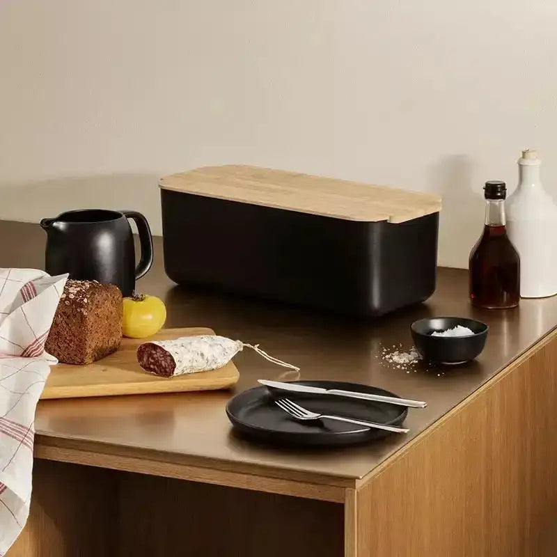Black bread bin with wooden lid on a kitchen counter with food items