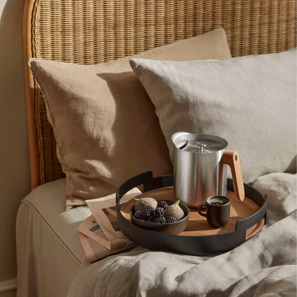 A styled bedroom scene with a bed made up in neutral beige linens, a woven rattan headboard, and a wooden tray holding a metal pitcher, a small dark cup, and a bowl of fruit. No person or animal is present, but this is not a clean isolated product shot; it is a real interior environment, so subject_type is Lifestyle.