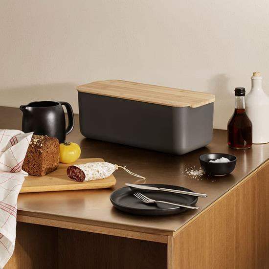 Black bread bin with wooden lid on a kitchen counter with bread and fruit