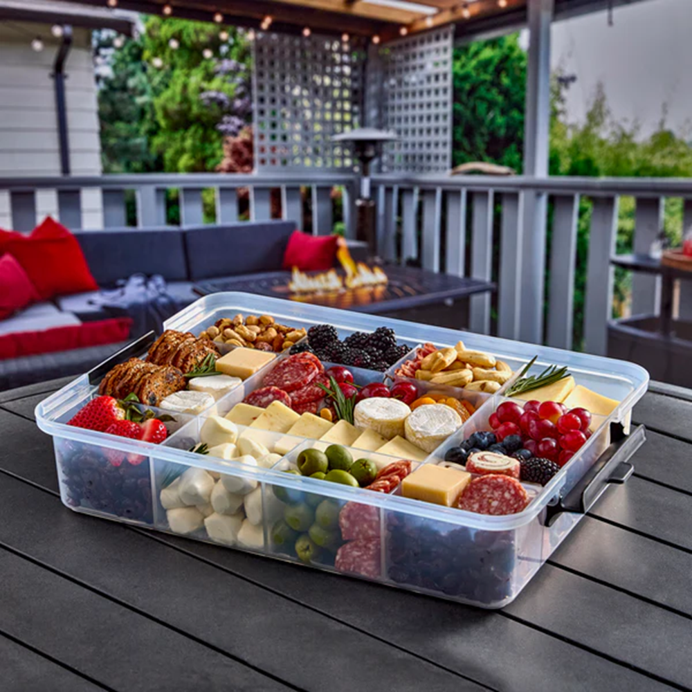 Assorted food platter in a clear container on a patio table with outdoor furniture in the background.