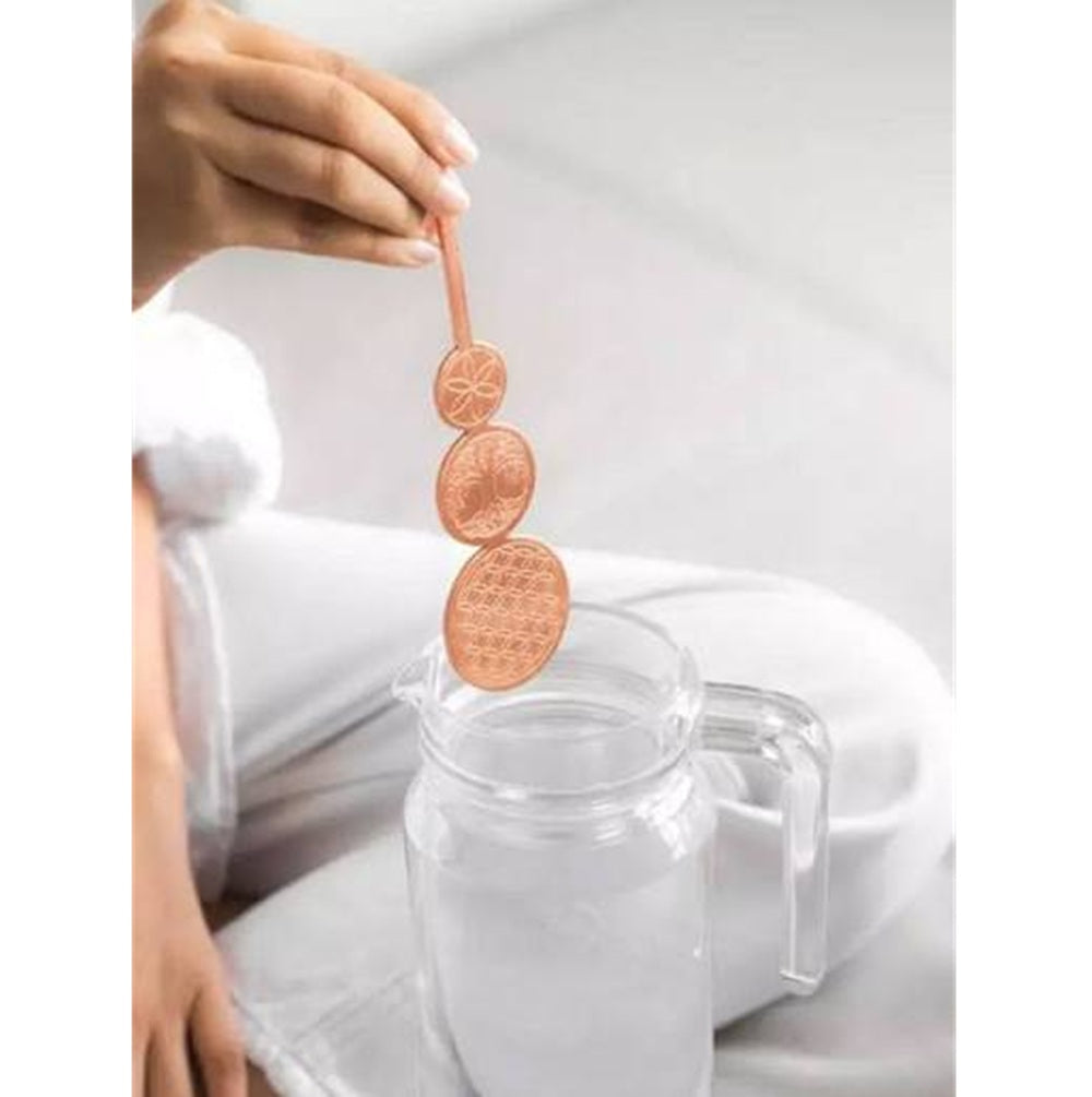 A person holds a copper-toned metal infuser with three circular sections above a clear glass pitcher filled with water.