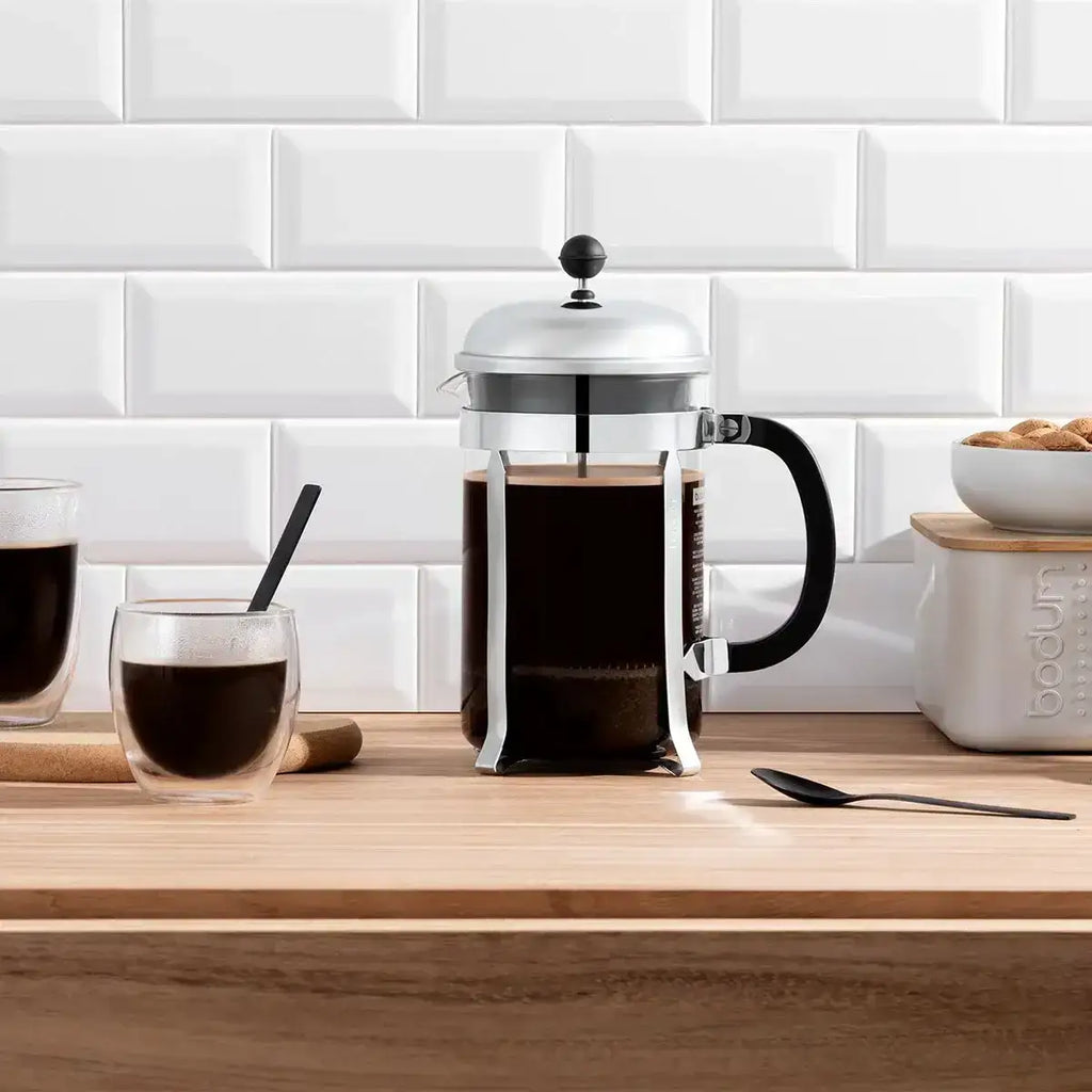 French press with coffee on a wooden counter against a white tiled wall.