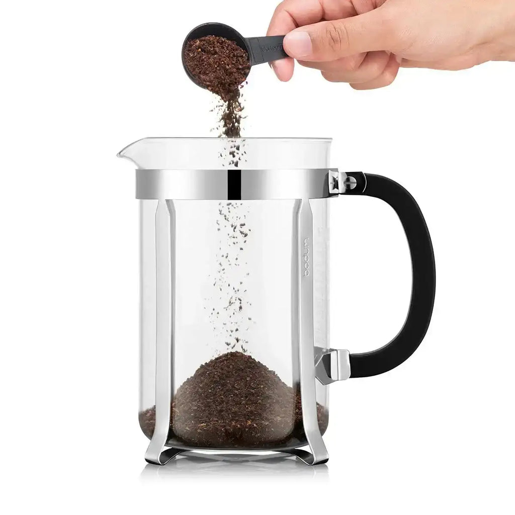 Hand pouring coffee grounds into a French press on a white background