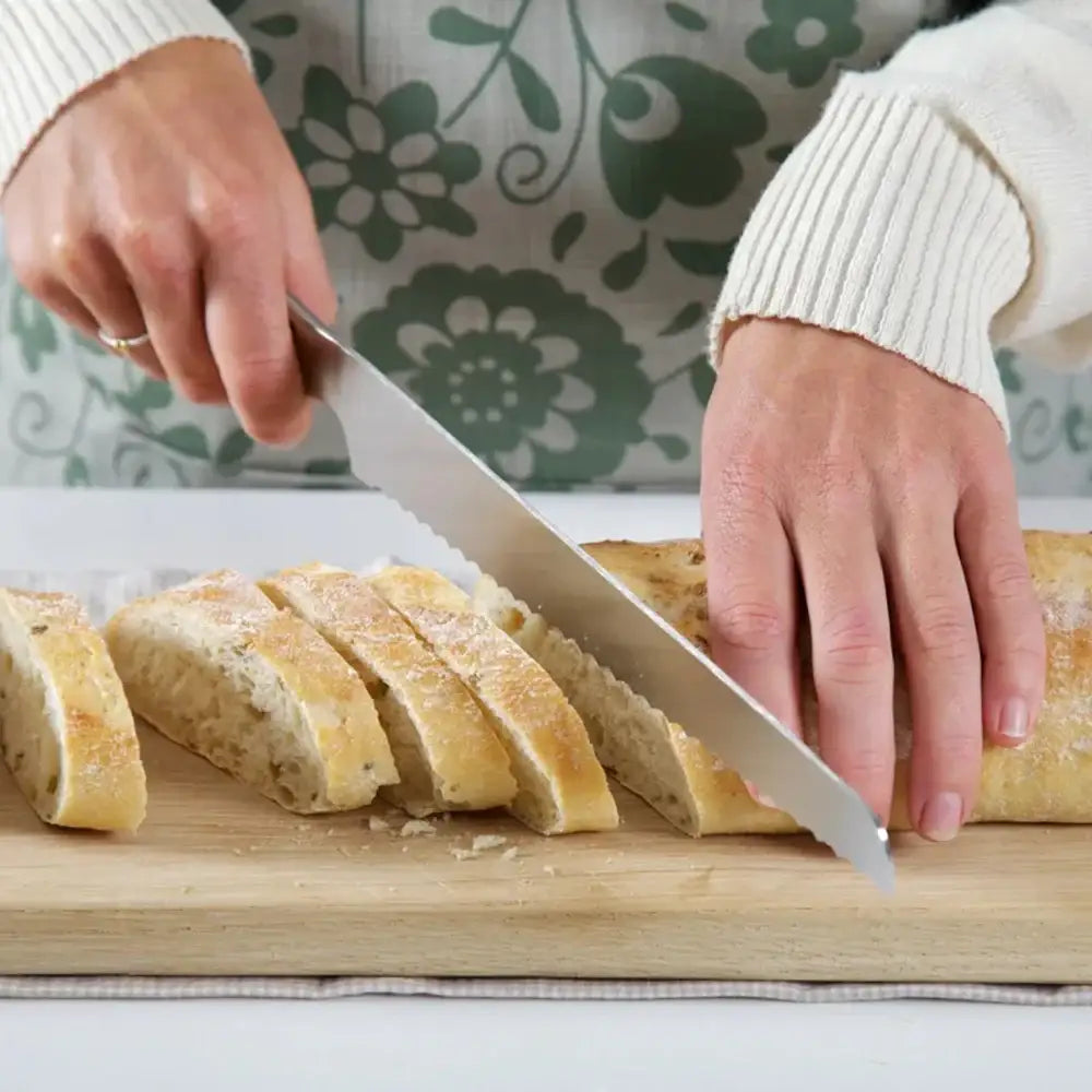 Person slicing bread on a wooden cutting board with a floral-patterned curtain in the background.