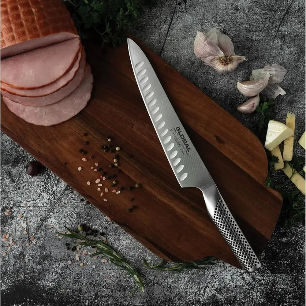 Chef's knife on a wooden cutting board with sliced ham, garlic, and herbs on a dark surface
