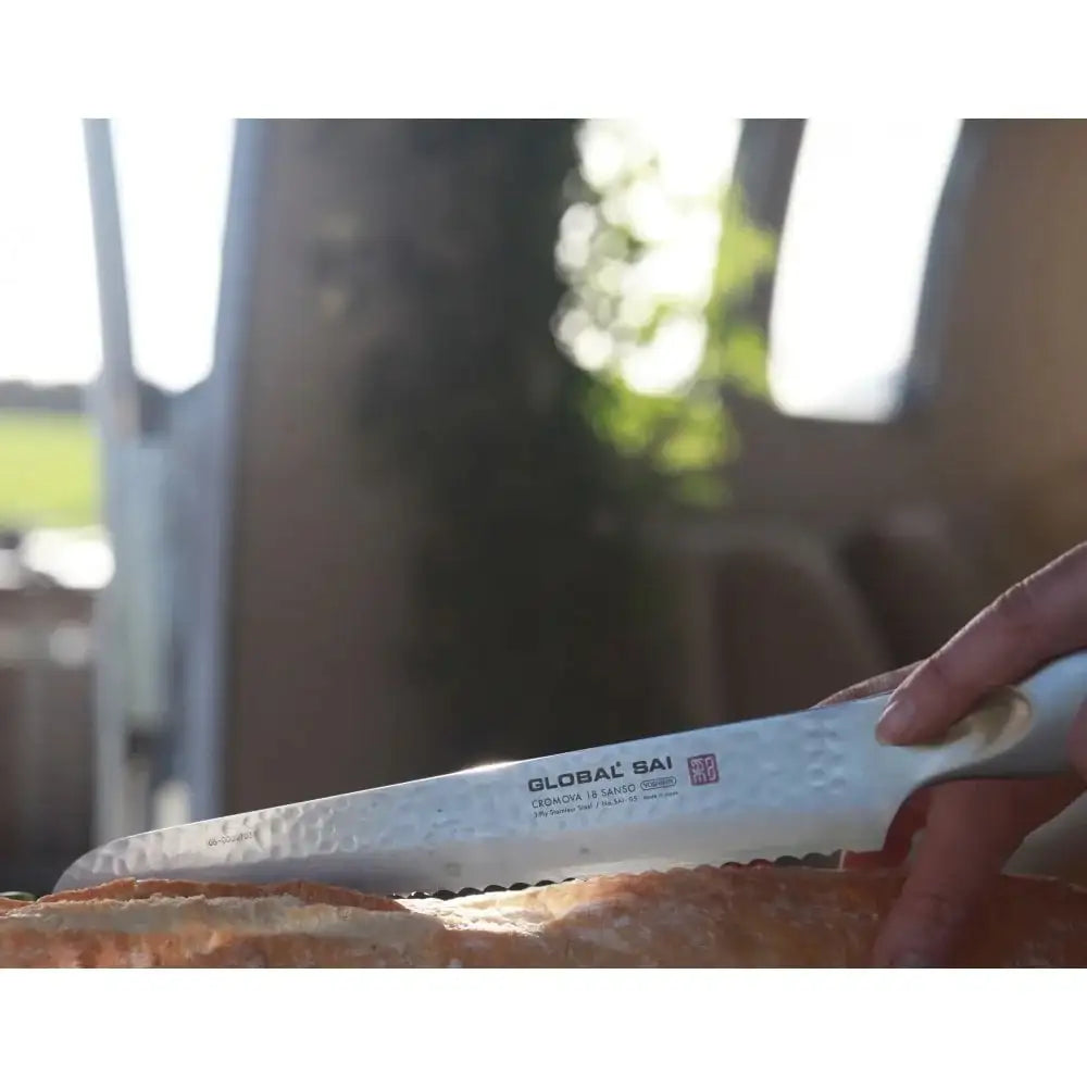 Person holding a bread knife with 'Global Sai' branding over a baguette.