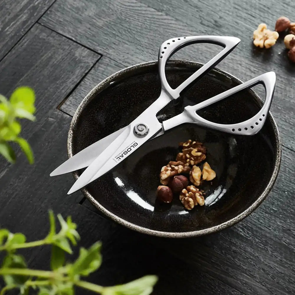 Pair of kitchen shears on a black plate with nuts and herbs on a dark surface