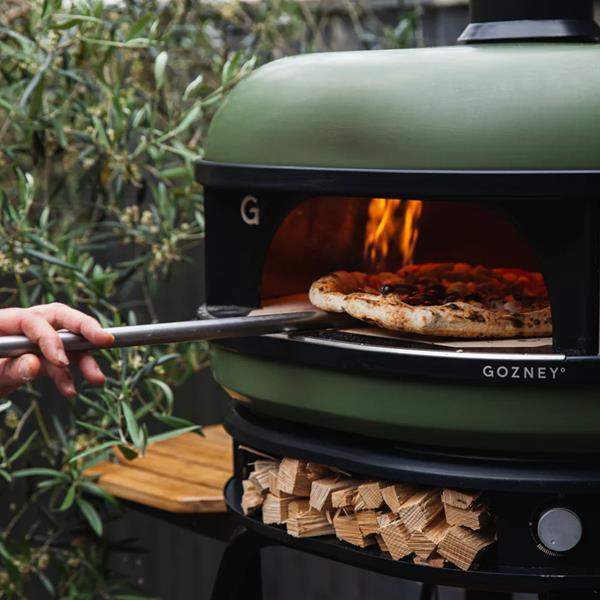 Outdoor Gozney Dome multi-fuel pizza oven baking pizza, shown with firewood at Cookin Stores.