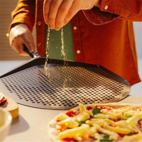 Person sprinkling flour on Gozney perforated pizza peel next to pizza, premium kitchenware at Cookin Stores