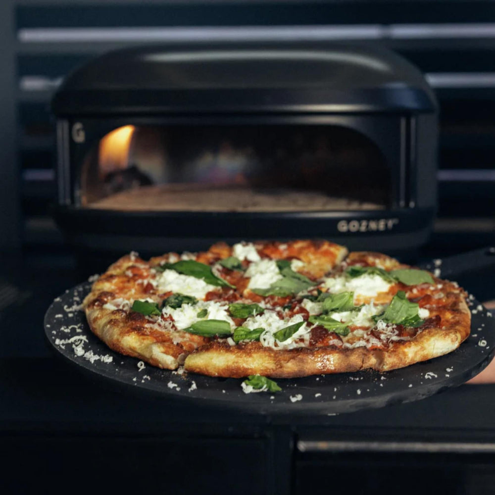 Pizza with green toppings on a black pan in front of a Gozney pizza oven.