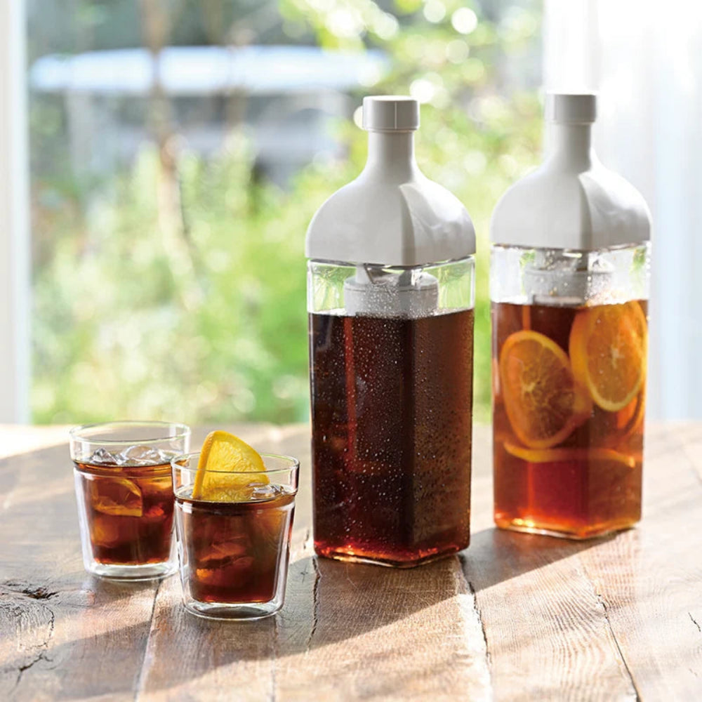 Two white glass soda bottles with clear lower bodies and two small clear glasses of iced cola on a wooden table, with citrus slices visible in the drink and a blurred outdoor background.