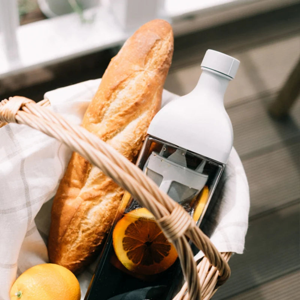 A white tea infuser bottle sits inside a woven wicker basket lined with white cloth, alongside a baguette, orange slices, and a whole orange.