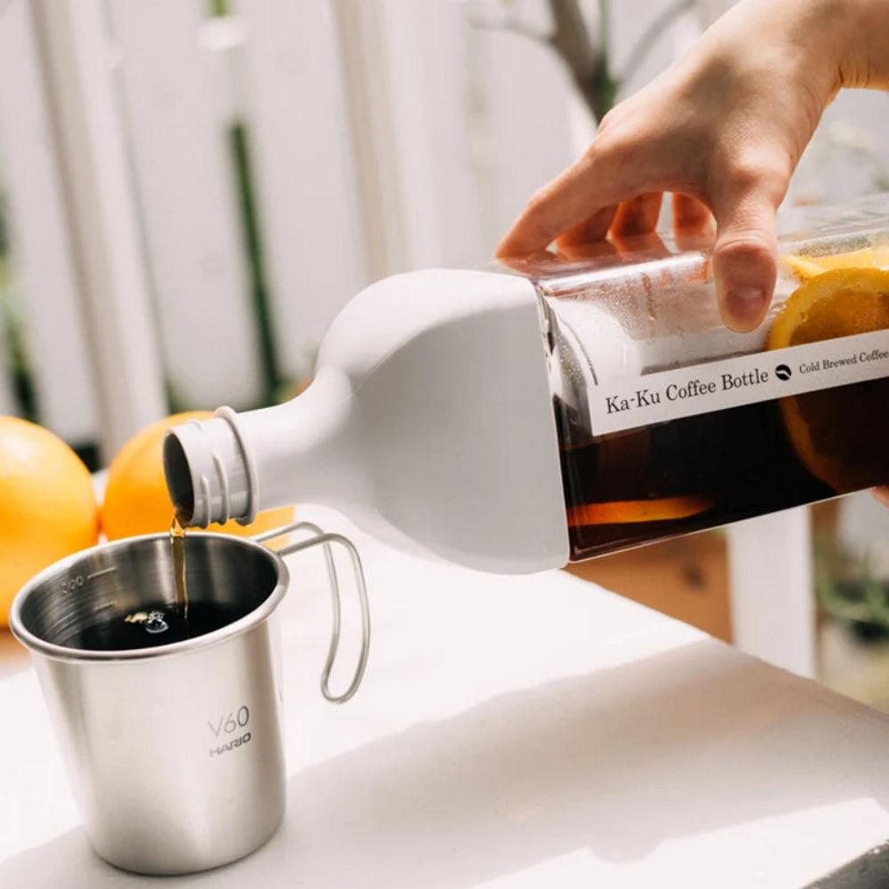A hand pours hot coffee from a white Ka-Ku Coffee Bottle with a clear glass body into a stainless steel mug. The bottle features a white body with a clear glass section showing amber coffee and a yellow citrus slice, and a black label with white text.