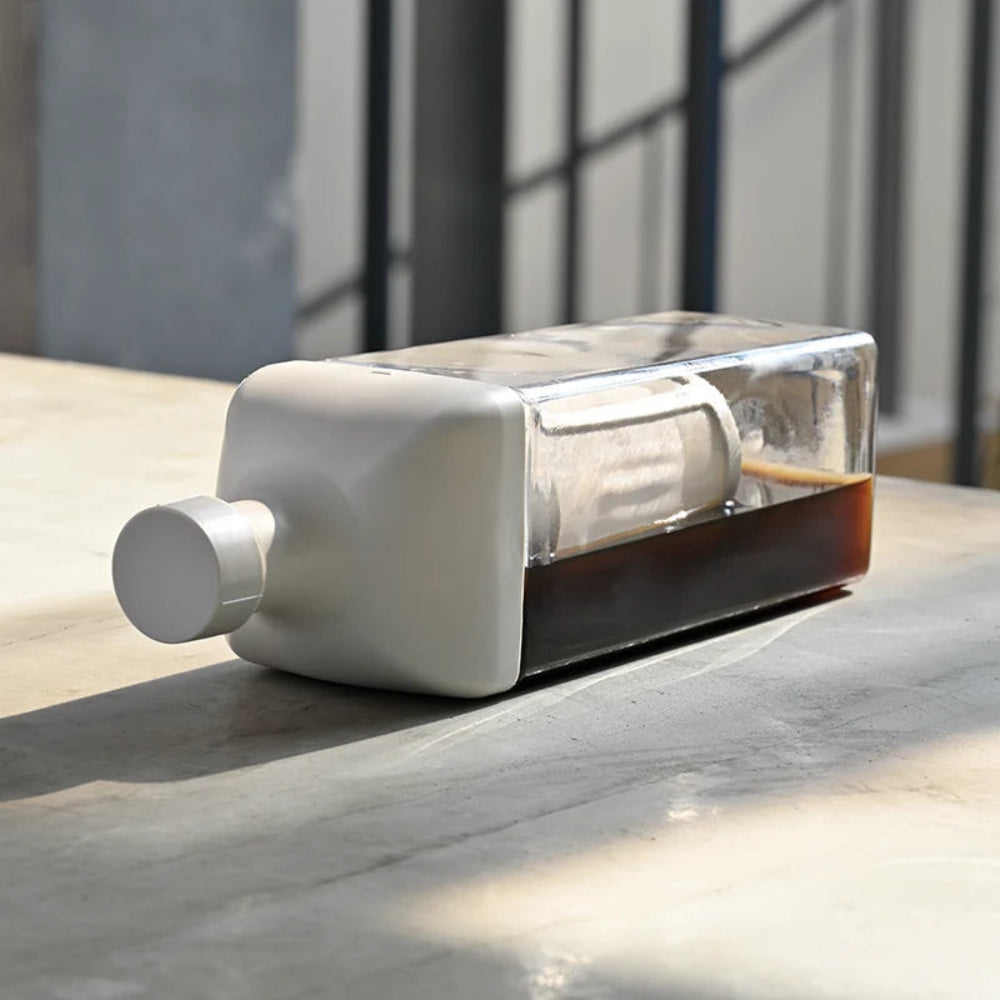 A modern rectangular bottle with a matte white body, a clear lid, and a white cylindrical filter or straw component, resting on a light stone surface with natural sunlight and shadows.