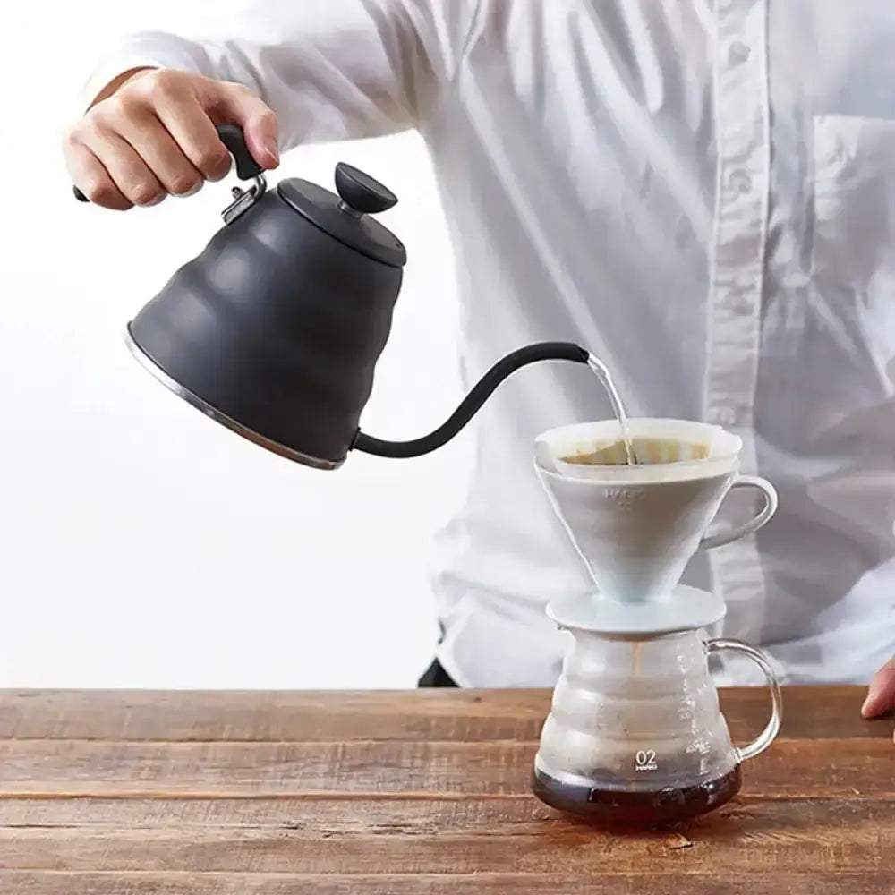 Person pouring coffee from a black kettle into a white coffee filter on a wooden table.