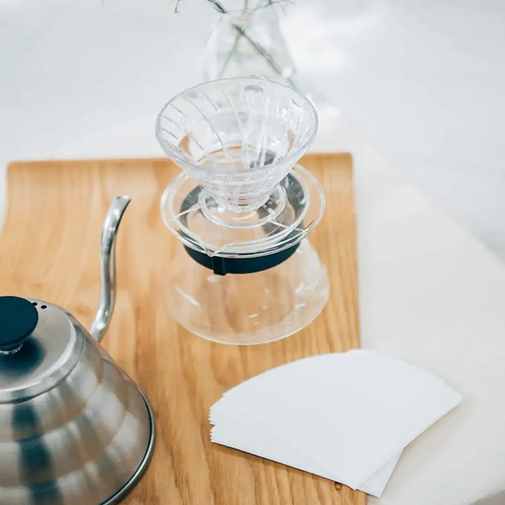 Clear glass coffee dripper on a wooden surface with a silver kettle and white paper filter.