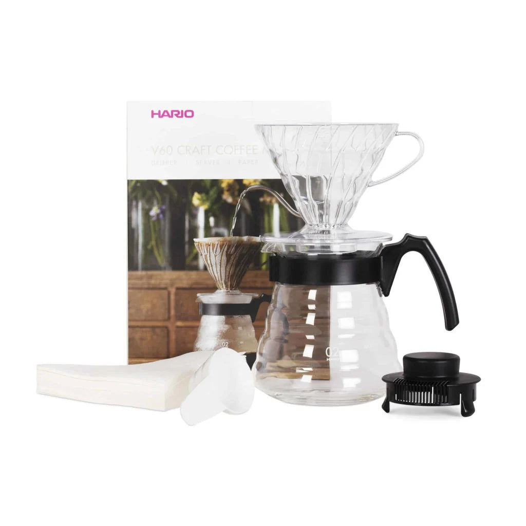 Hario coffee brewing equipment including a clear glass carafe with black lid and handle, white filter, and black scoop on a white background.