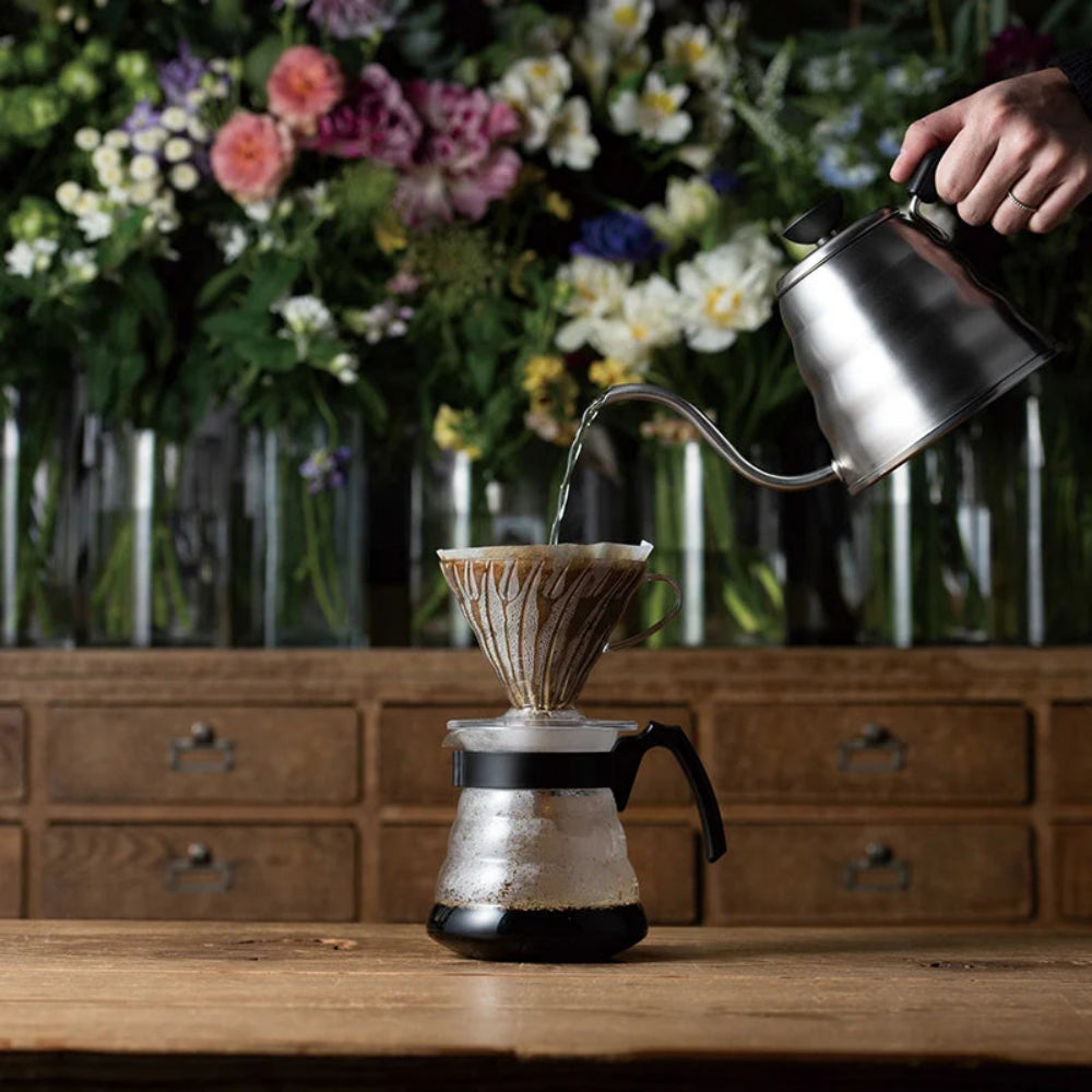 Person pouring water from a kettle into a coffee filter on a wooden table with flowers in the background