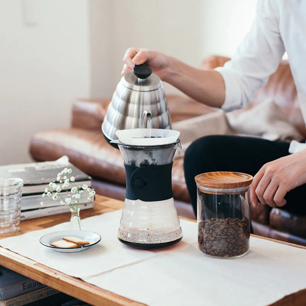 Person making coffee using a manual coffee press on a table with a cozy living room background.