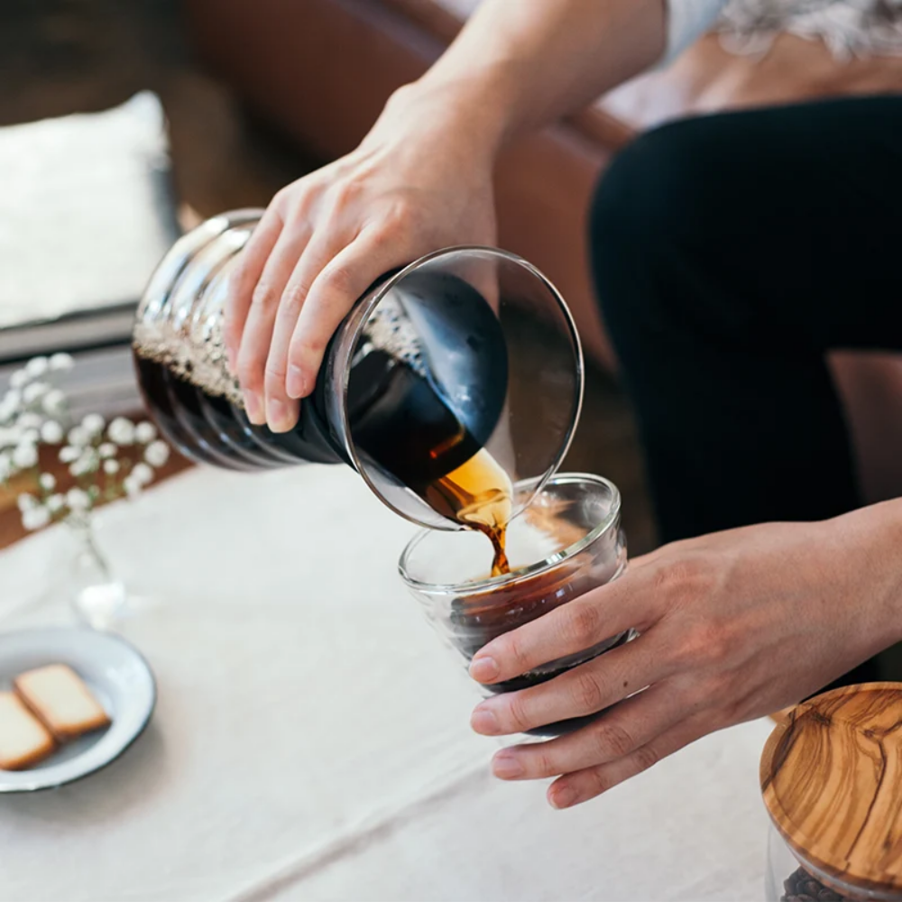 Person pouring coffee from a carafe into a glass on a table.