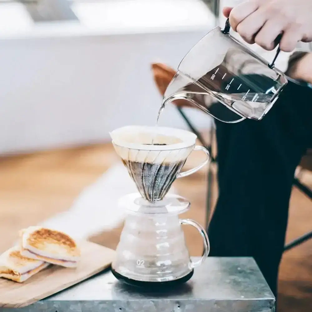 Person pouring liquid from a clear glass pitcher into a coffee filter on a wooden table.