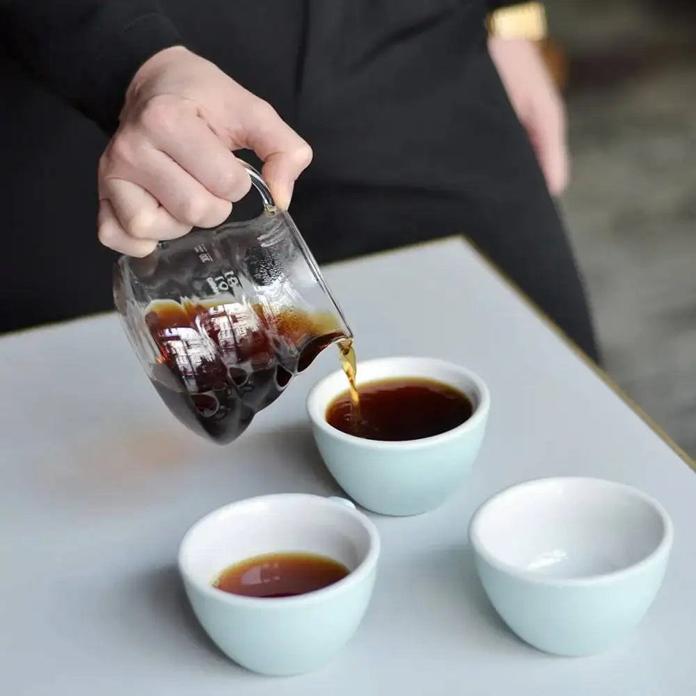 Person pouring dark liquid from a glass container into three small white cups on a light surface.