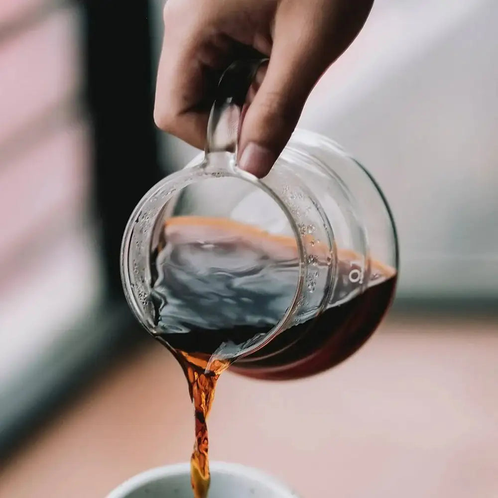 Hand pouring coffee from a glass carafe into a white cup.