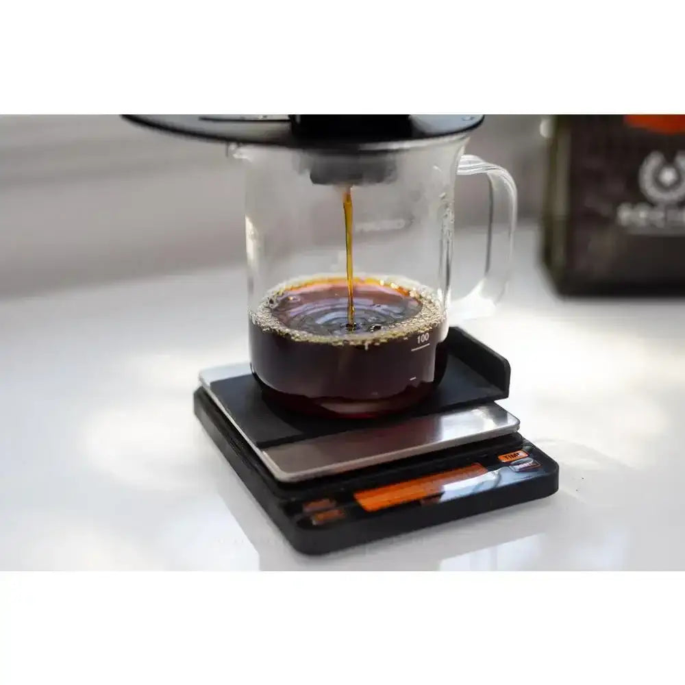 Coffee being poured into a glass carafe on a digital scale with a coffee bag in the background.