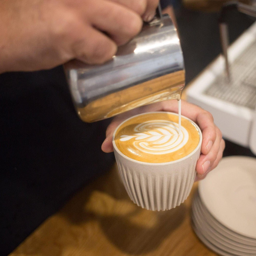 Person pouring a latte into a white cup with a decorative pattern.