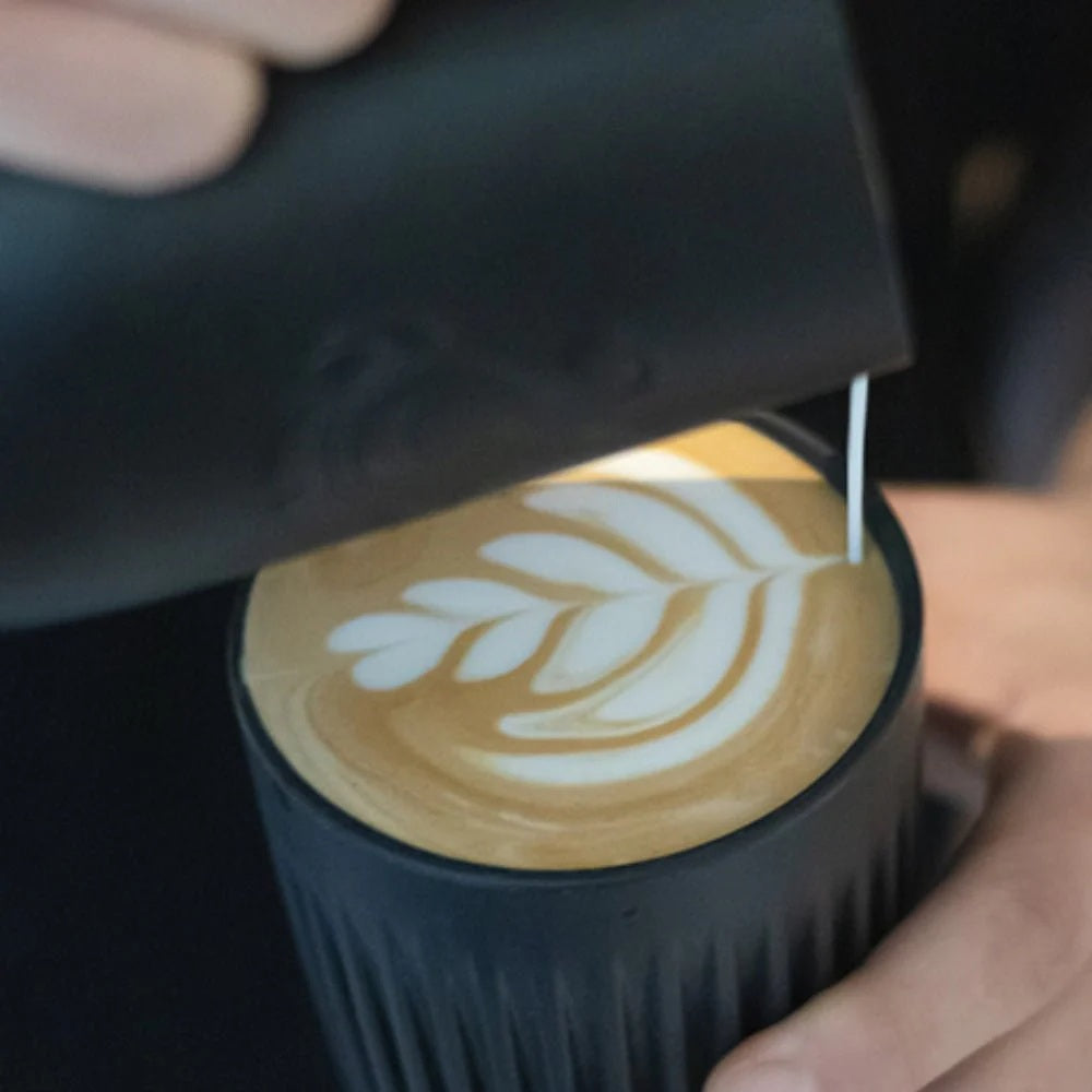 Person pouring a dark liquid into a cup with latte art, held by another hand.