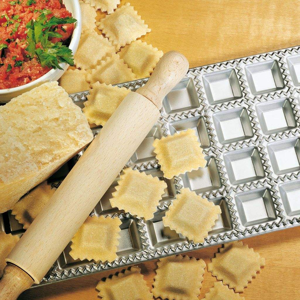 Ravioli making setup with a rolling pin, pasta press, and ravioli on a wooden surface.