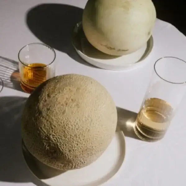Two cantaloupes on plates with glasses of liquid on a white surface