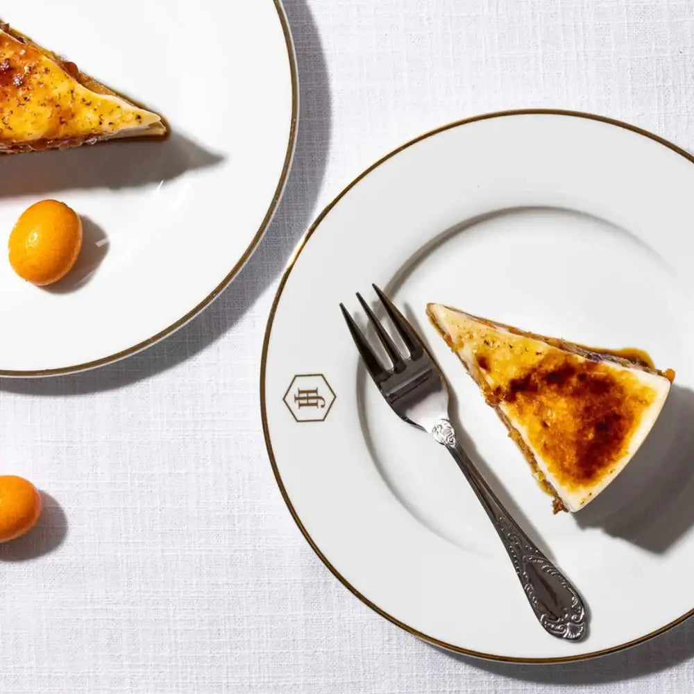 Slice of creme brèleé on a plate with a fork, surrounded by oranges on a white tablecloth.