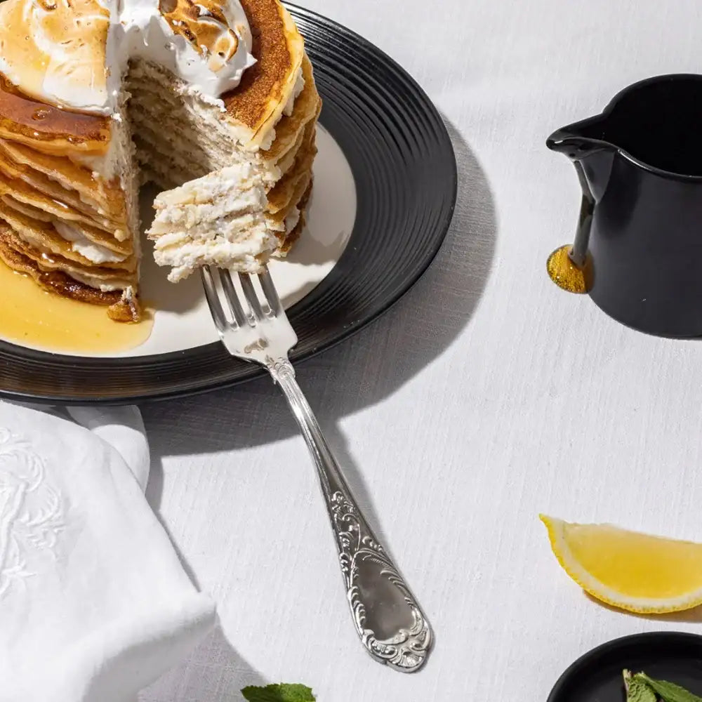 Stack of pancakes with a slice cut out, served on a black plate with a fork, on a white tablecloth.