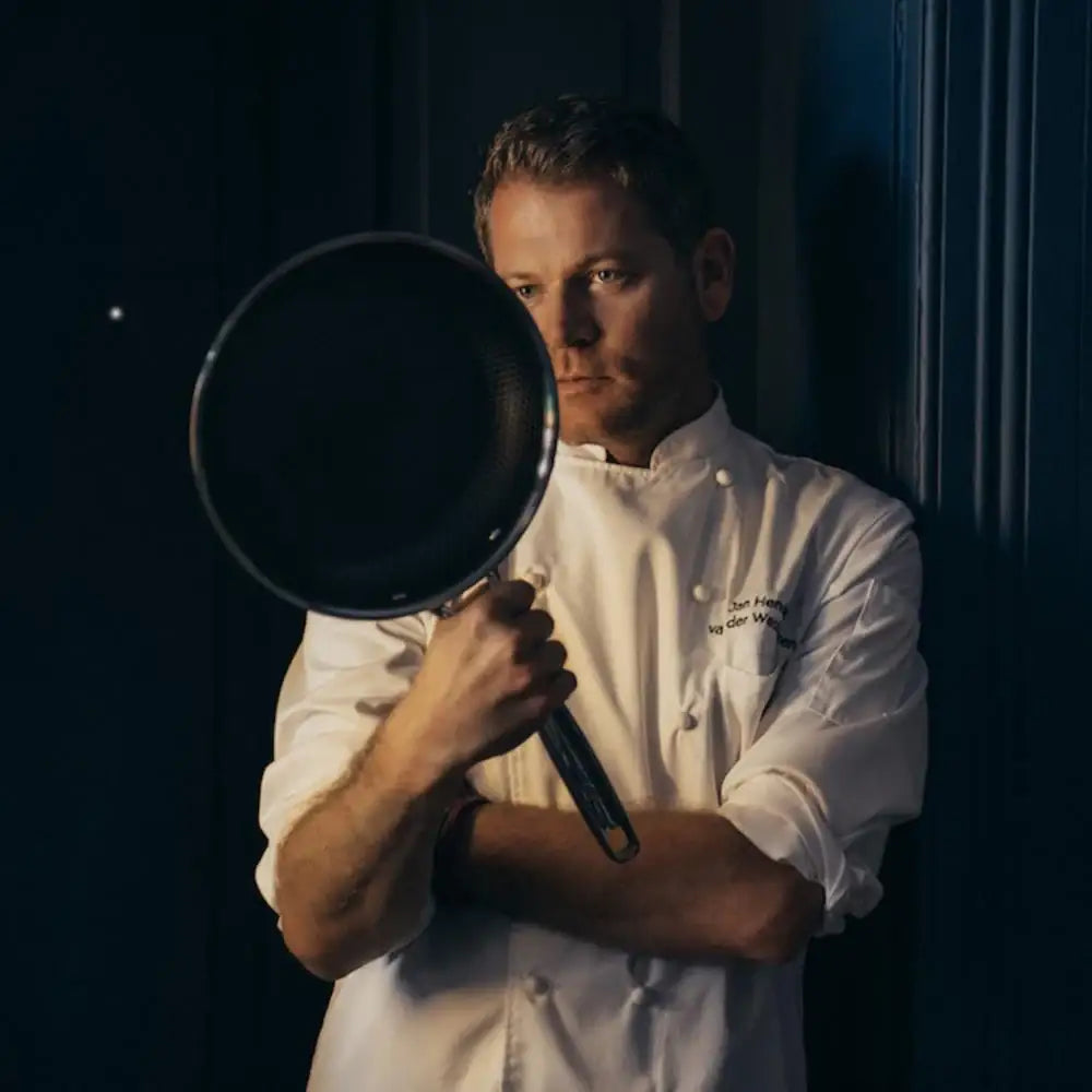 Man in a chef's coat holding a frying pan against a dark background