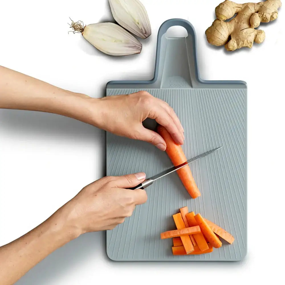 Person cutting carrots on a gray cutting board with onions and ginger in the background.