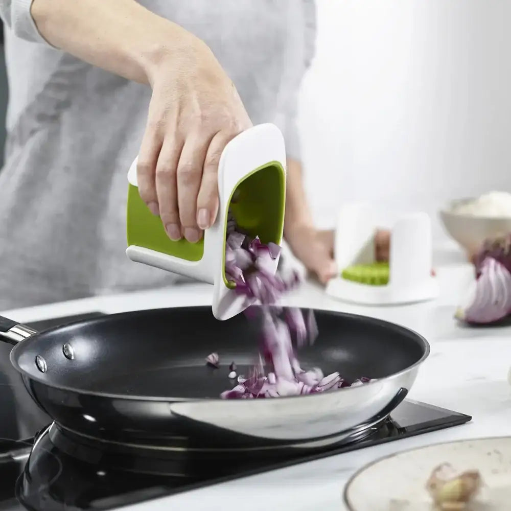 Person using a green and white kitchen tool to add chopped onions into a black frying pan on a stove.