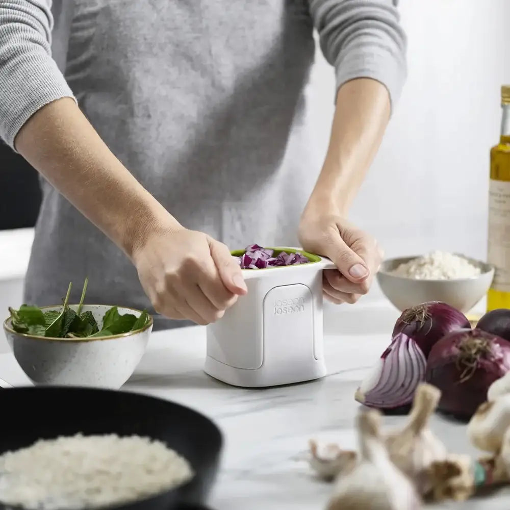 Person using a kitchen tool to chop vegetables on a counter with various ingredients around.