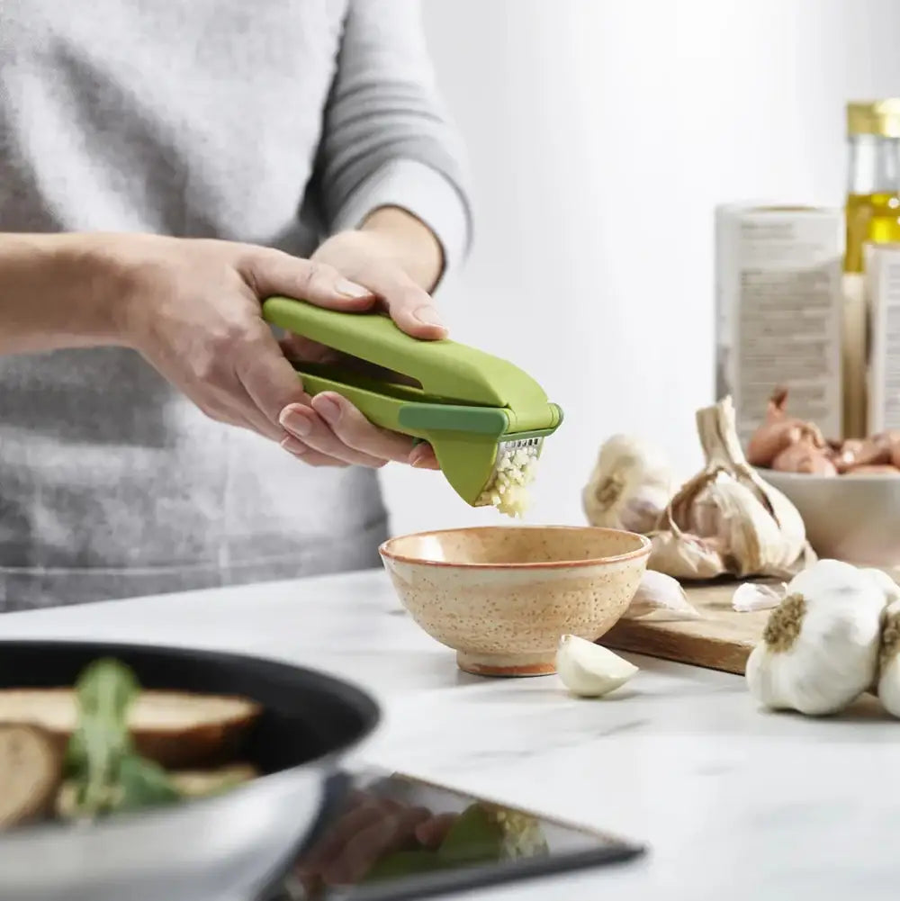 Person using a green garlic press on a kitchen counter with garlic and a bowl.