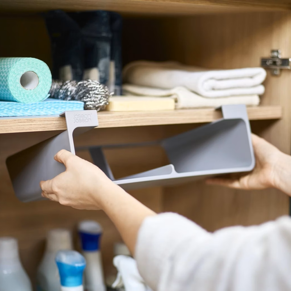 A person's hands sliding a gray plastic under-cabinet organizer tray into a wooden bathroom or utility cabinet, with toiletries visible on the shelf.