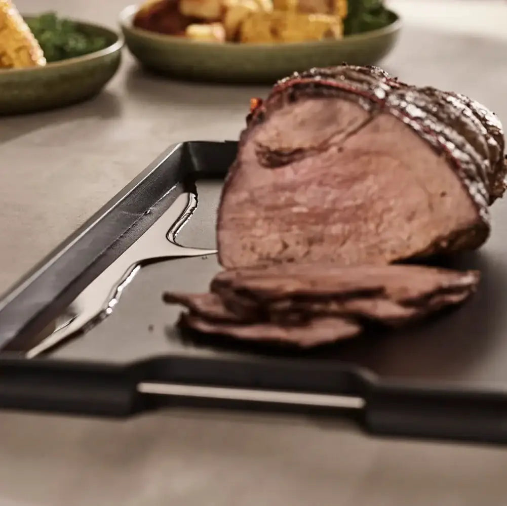 Sliced roast beef on a cutting board with a knife and fork, surrounded by other dishes.