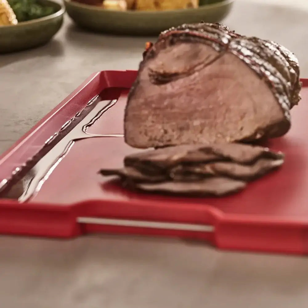 Sliced roast beef on a red cutting board with a blurred background of a dining table.