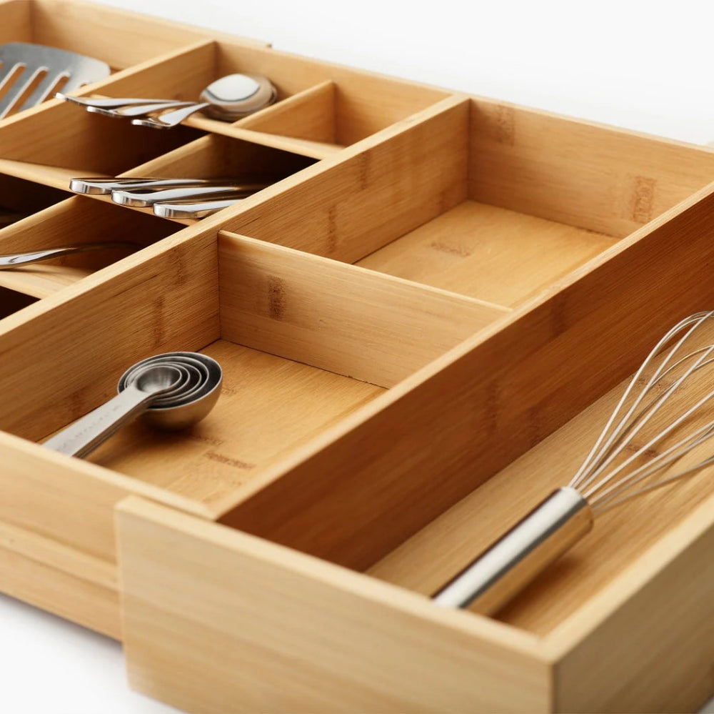 A bamboo kitchen utensil organizer drawer with multiple compartments holding metal measuring cups, a whisk, and other kitchen tools on a white background.