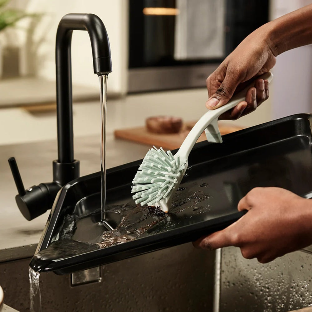 Person cleaning a black tray with a white scrubbing brush in a kitchen.