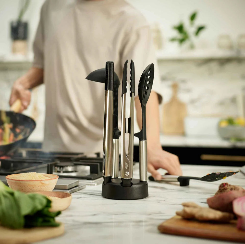 A stainless steel silicone utensil kitchen set on a marble countertop, with a person cooking in a blurred kitchen background.