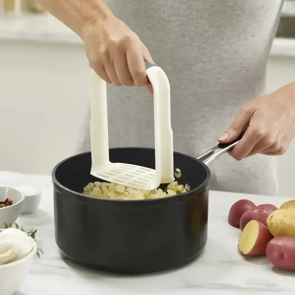 Person mashing potatoes in a pot with a white masher, surrounded by ingredients on a kitchen counter.