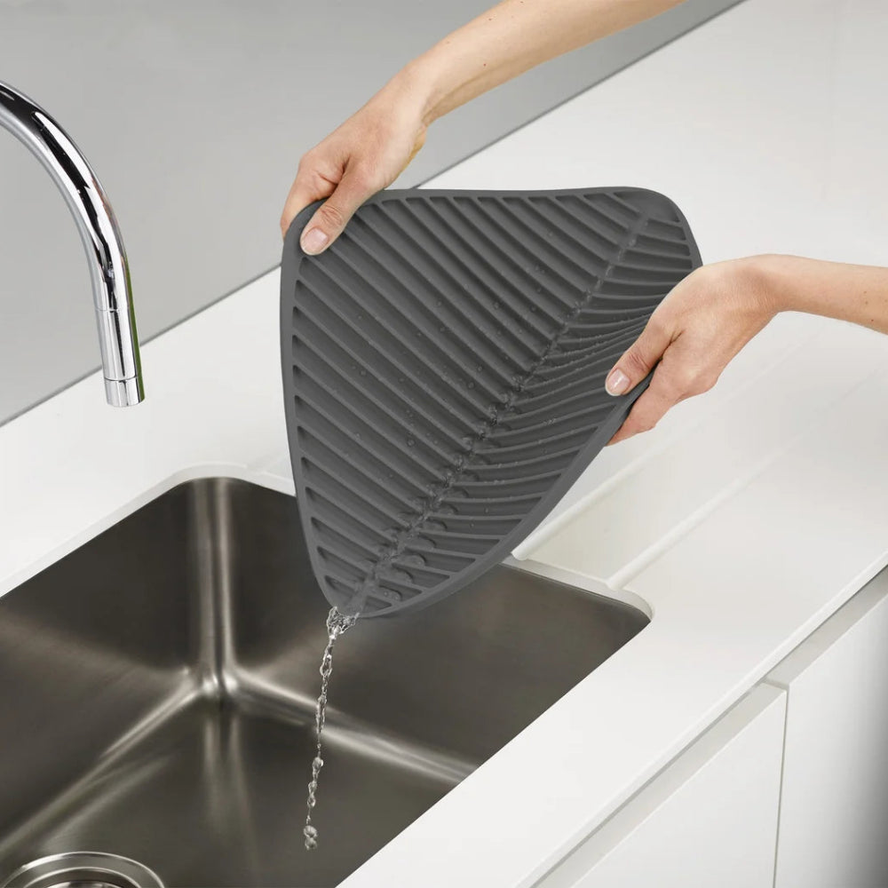 A person holds a dark gray silicone kitchen mat with a leaf-like shape over a stainless steel sink, with water dripping from the edge, in a bright modern kitchen setting.