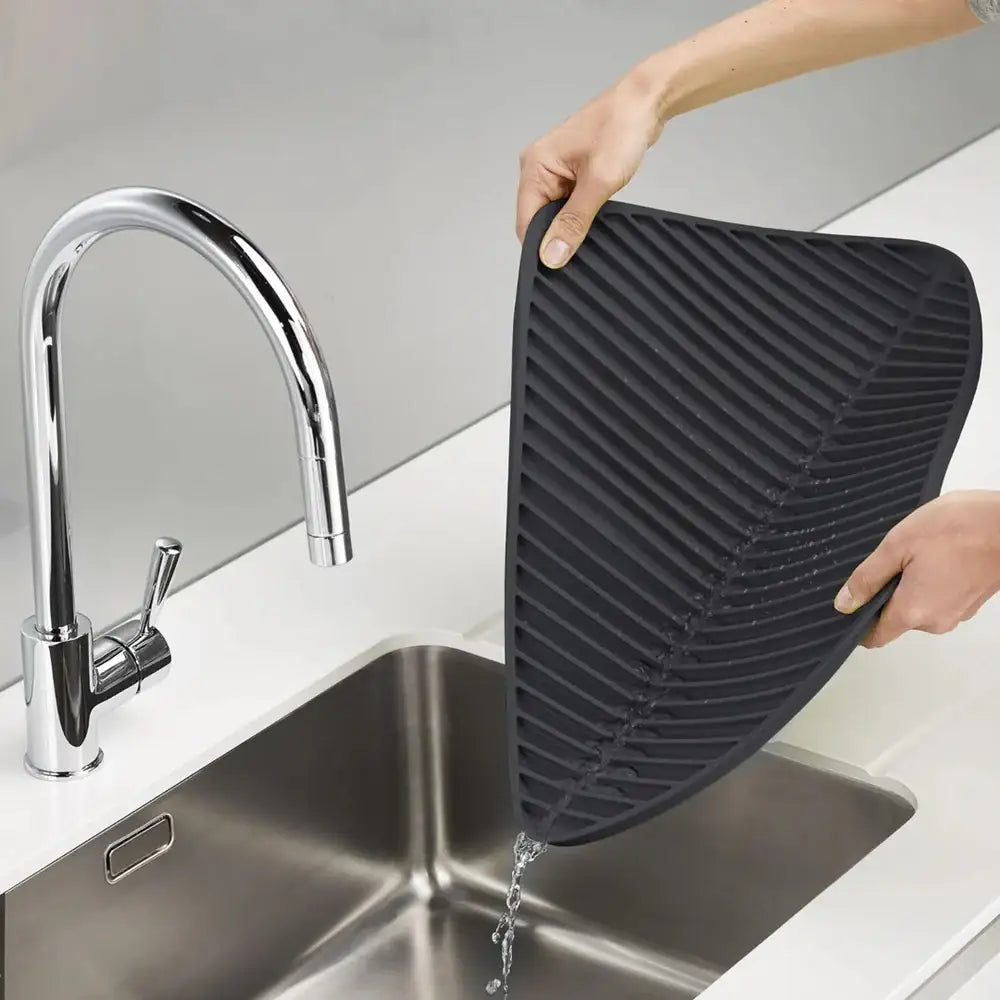 Person holding a black silicone mat over a kitchen sink with water running.