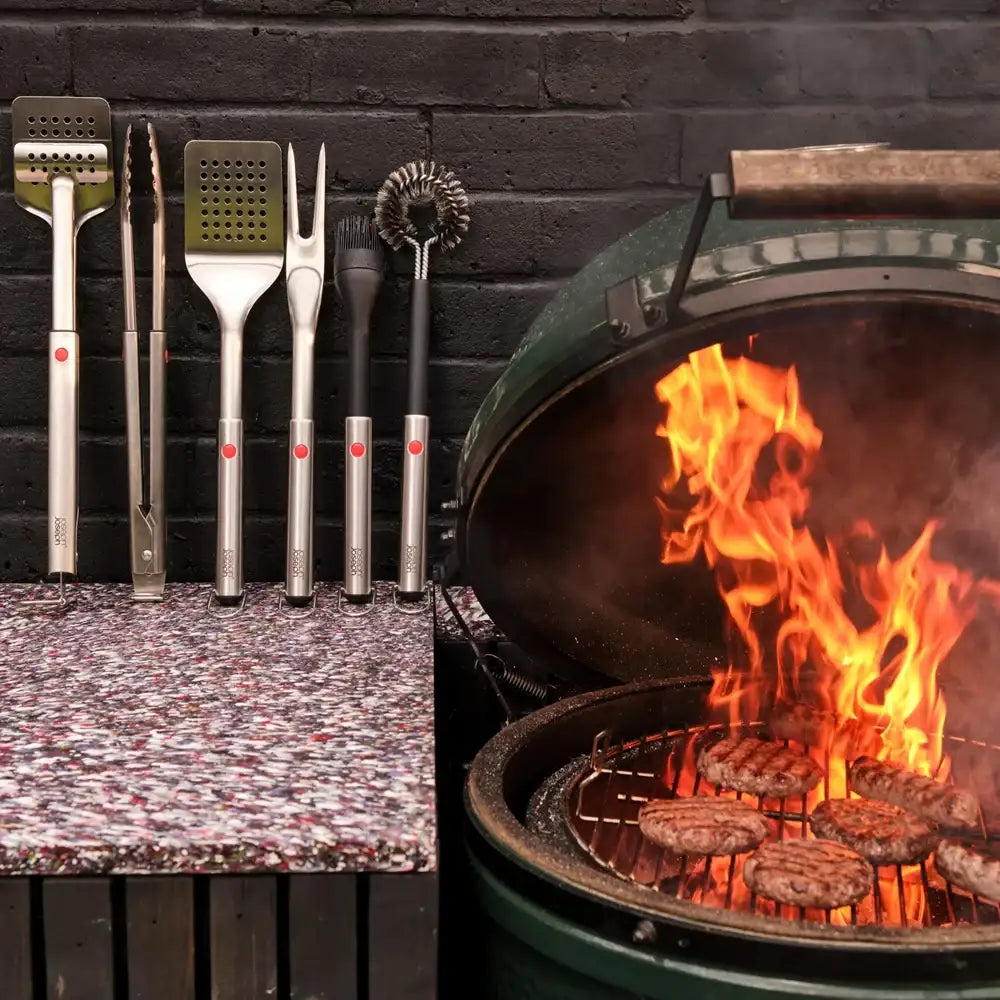 Barbecue grill with flames and burgers cooking, alongside a set of grilling tools on a stone surface.