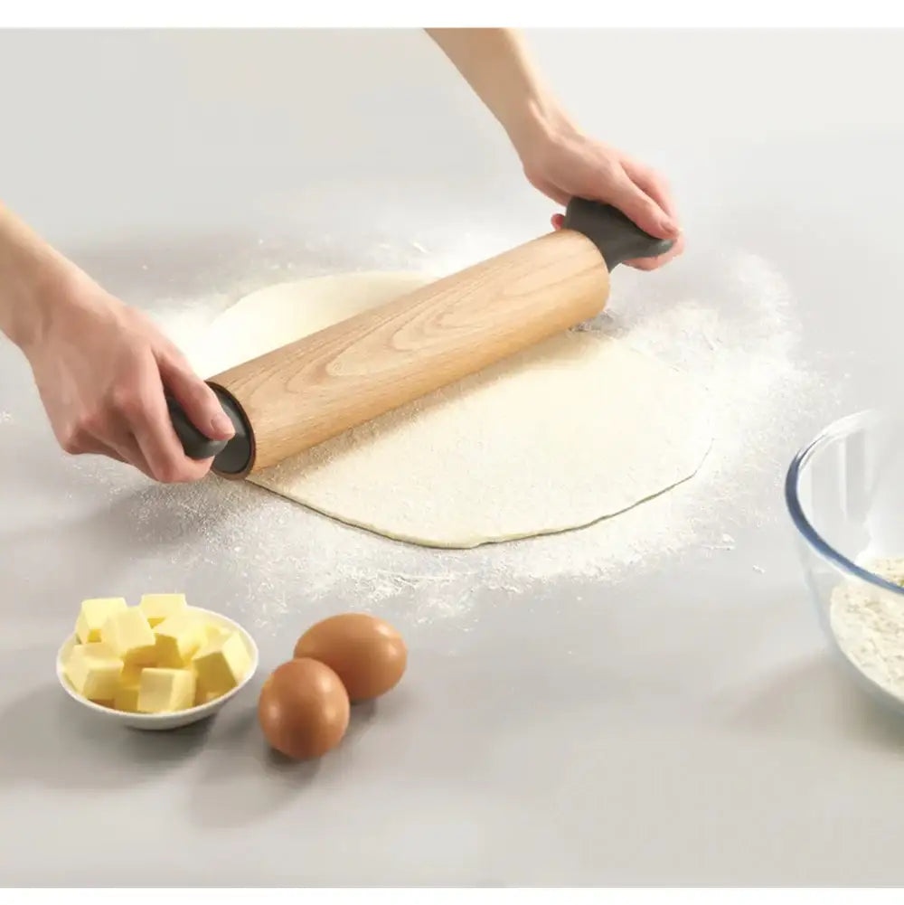 Person rolling out dough with a wooden rolling pin on a light surface, with butter and eggs nearby.
